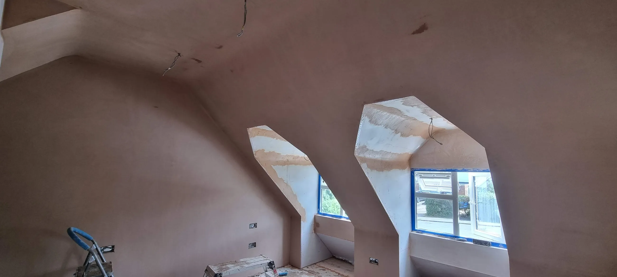 A freshly plastered loft room with two dormer windows set into the sloping ceiling, the plaster still pink and drying.