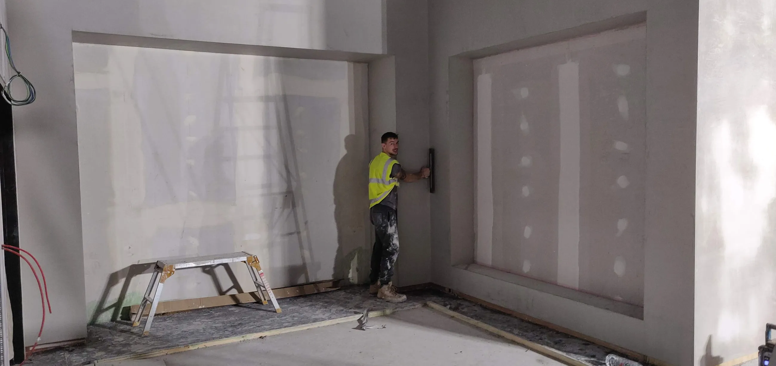 An interior room being skimmed, with a plasterer working a wall behind protective sheeting.