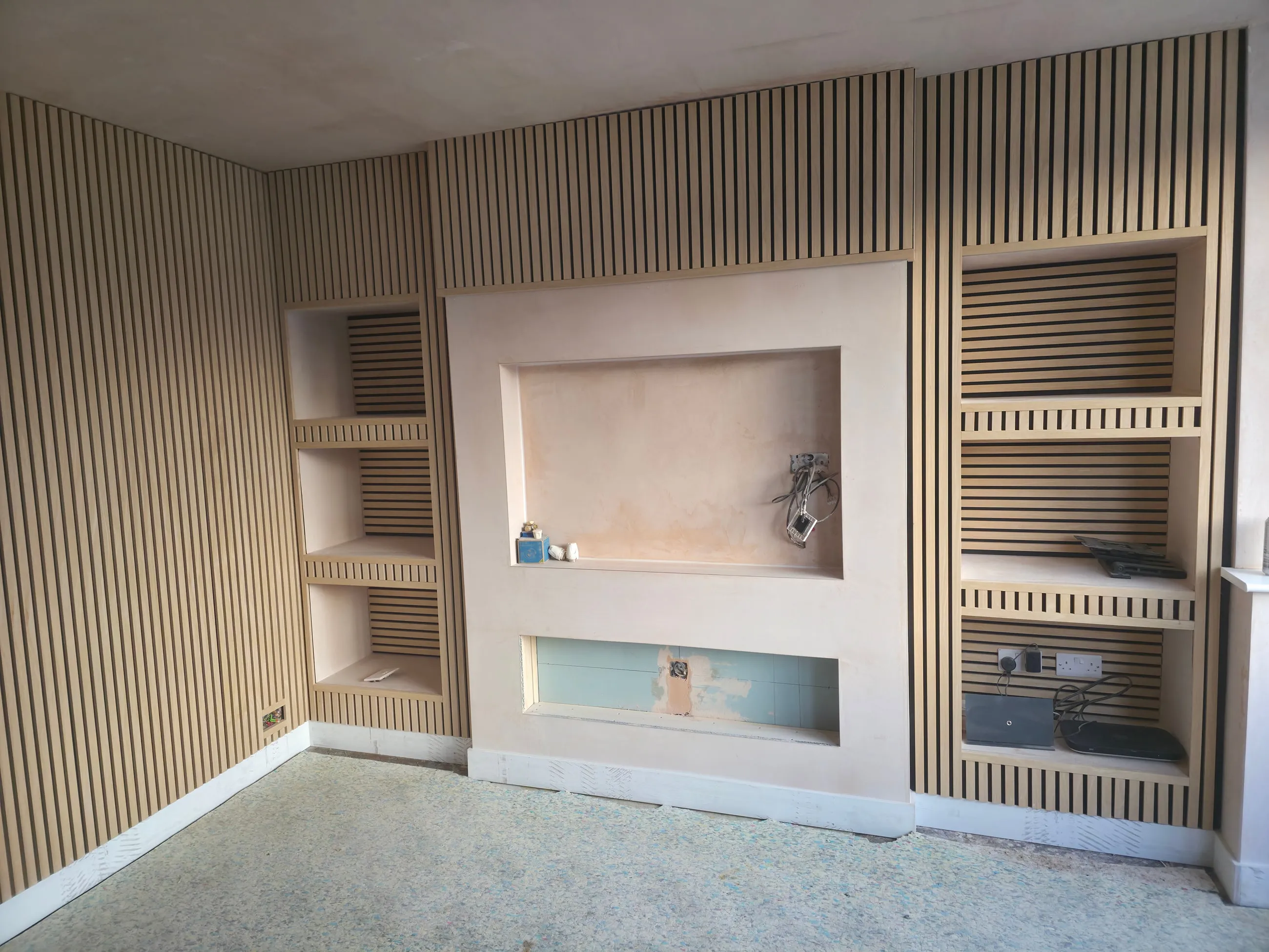A finished lounge media wall: slatted timber panelling across the wall, with a large plastered recess for a television and built-in shelving alcoves either side.