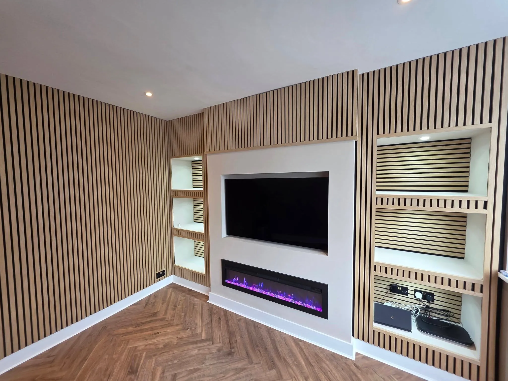 The finished media wall in oak slatted panelling, with a recessed television above a linear electric fire and lit display alcoves either side.