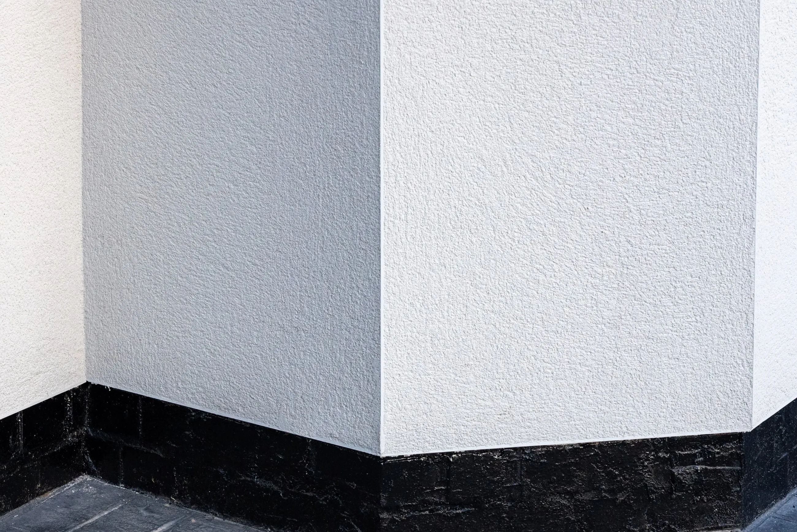 A close view of a rendered external corner: crisp white textured render meeting a dark plinth in a clean, straight line.