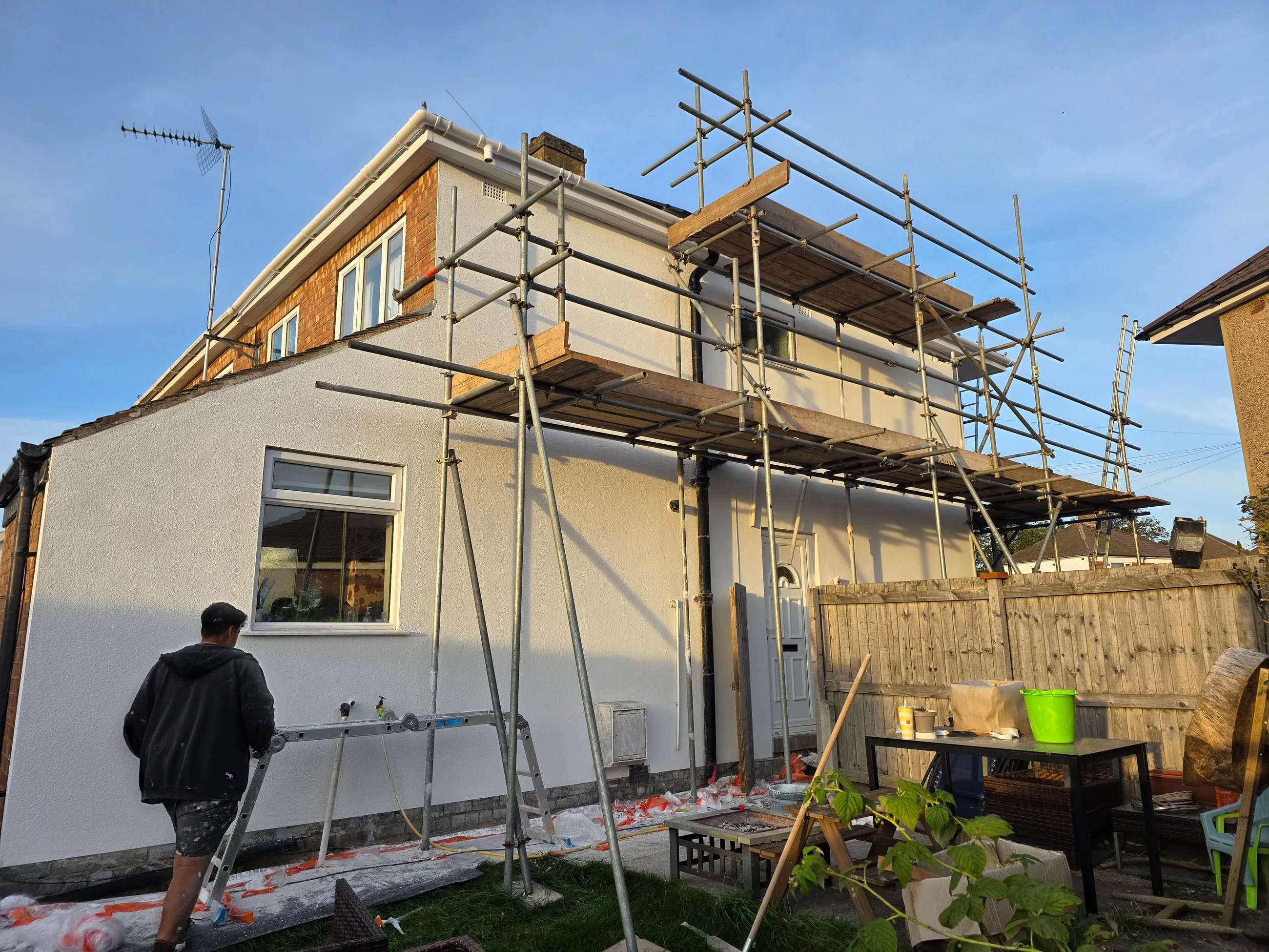 A house being rendered, one elevation finished in white and the scaffold still standing, with a member of the team working at the base.