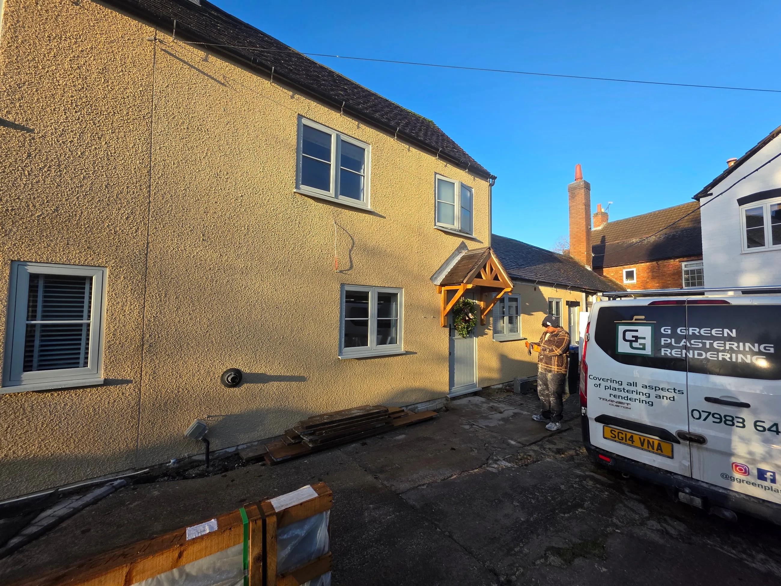 A cottage finished in heavy-texture cream roughcast, lit by low winter sun, with sage-green windows and an oak porch. One of the team is working at the door and the G Green van is parked alongside.