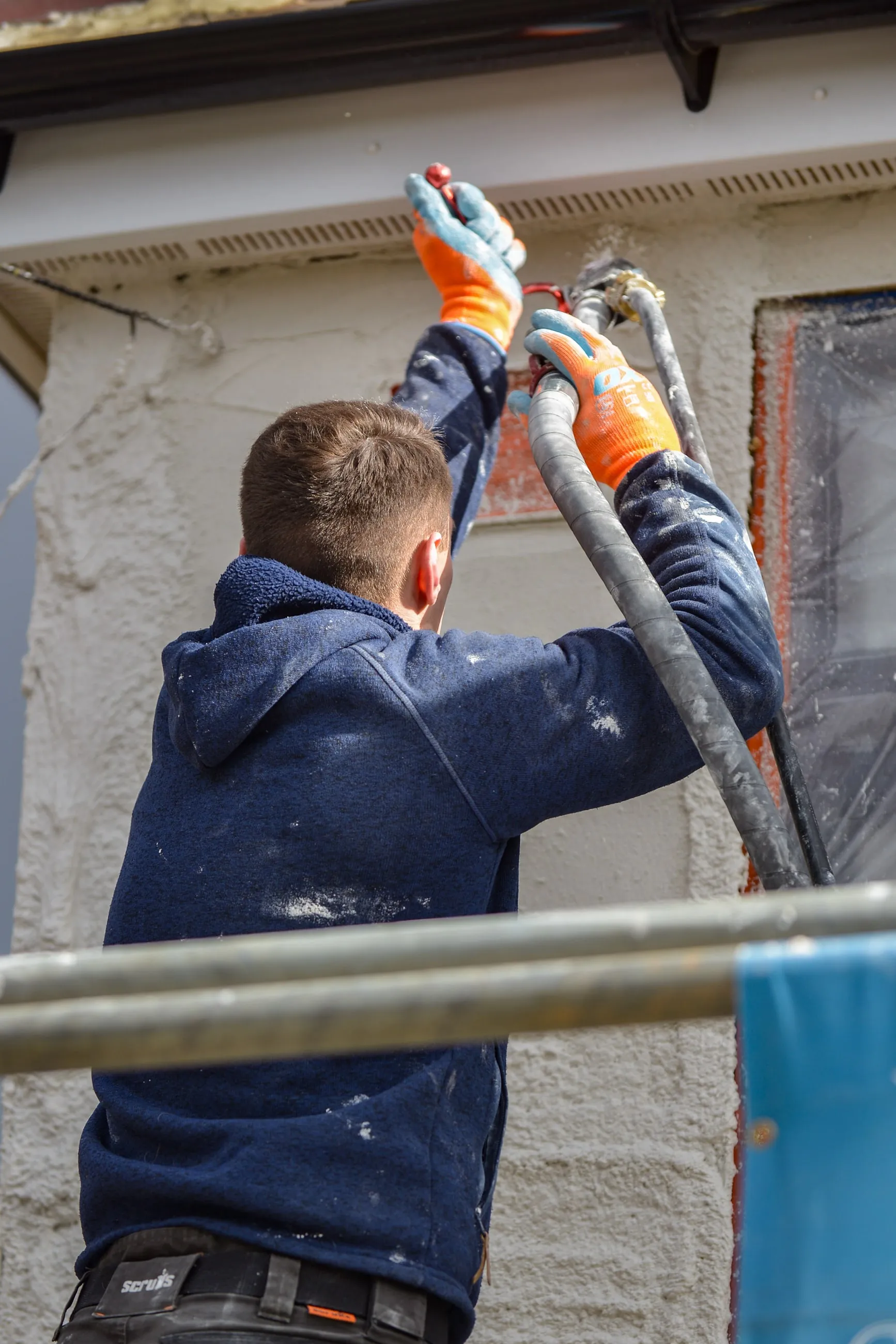 A member of the team on a scaffold, spraying monocouche render onto a wall through a hose held above his head.